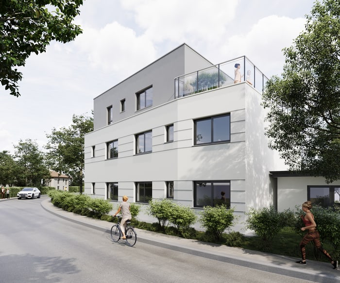 Modernes Wohngebäude mit mehreren Etagen, Balkon und Dachterrasse, umgeben von Bäumen und einer Straße.