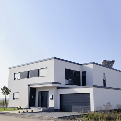 Modernes, weißes Haus mit Flachdach, Garage, dunklen Akzenten und Solaranlage unter klarem Himmel.